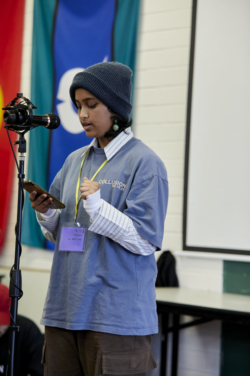 Image of a young person wearing a beanie and a blue top over a shirt, speaking to a microphone and holding a phone