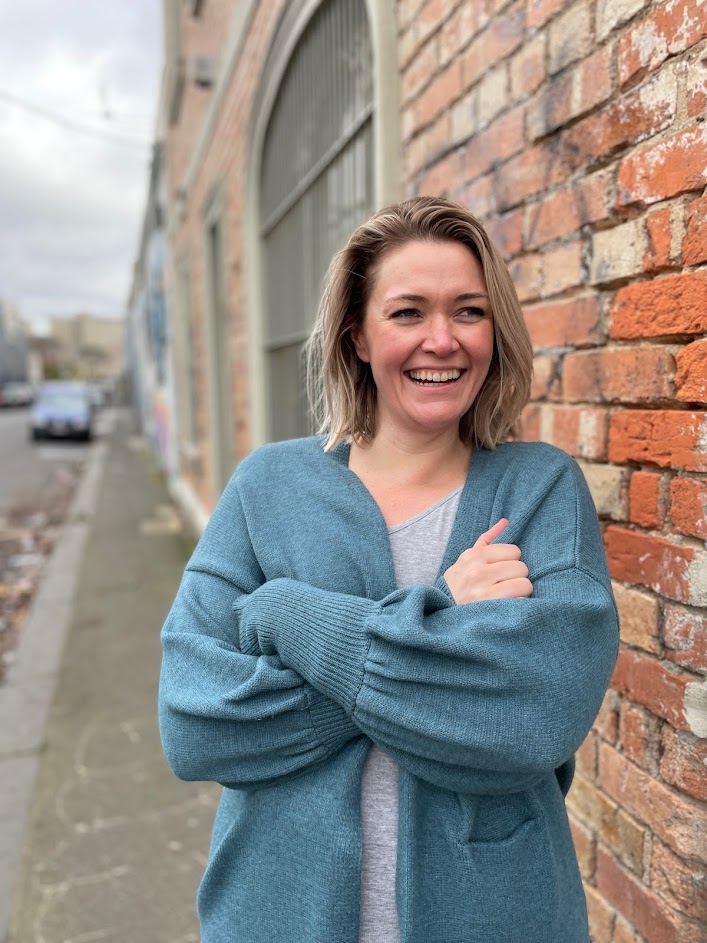Lauren is wearing a pale blue cardigan with a grey t-shirt underneath. She is standing next to a brick wall, with her arms crossed, smiling at the camera.