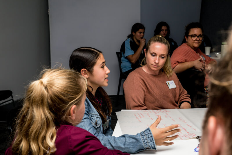 Young person with dark long hair and a denim jacket, talking amongst a group of people on a table at the Young Mayors Mackay Co-design workshop