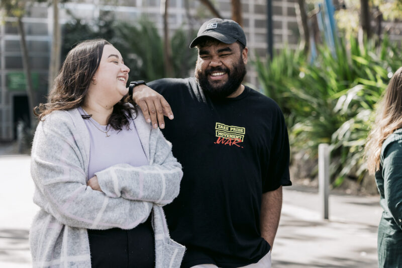 Picture of two First Nations People walking and smiling at each other. On the left is a woman wearing a grey shirt and jumper with her hands crossed. On the right is a man wearing a black cap and shirt, with his hands leaning on the woman's shoulder.