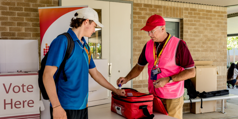Student wearing a blue uniform and white cap has his hand placed on a red ballot box. A person wearing a pink fluorescent cap and red cap is placing a purple ink stamp on the student's hand.